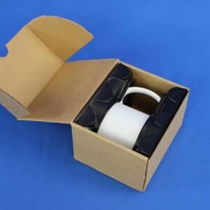 May include: A white ceramic mug is inside a brown cardboard box with a black foam insert. The box is open and the mug is visible.