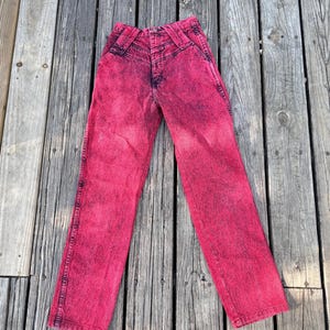 May include: A pair of acid wash pink denim jeans. The jeans have a high waist and a classic five-pocket design. The denim is a vibrant pink color with darker pink accents, giving it a unique, vintage look.