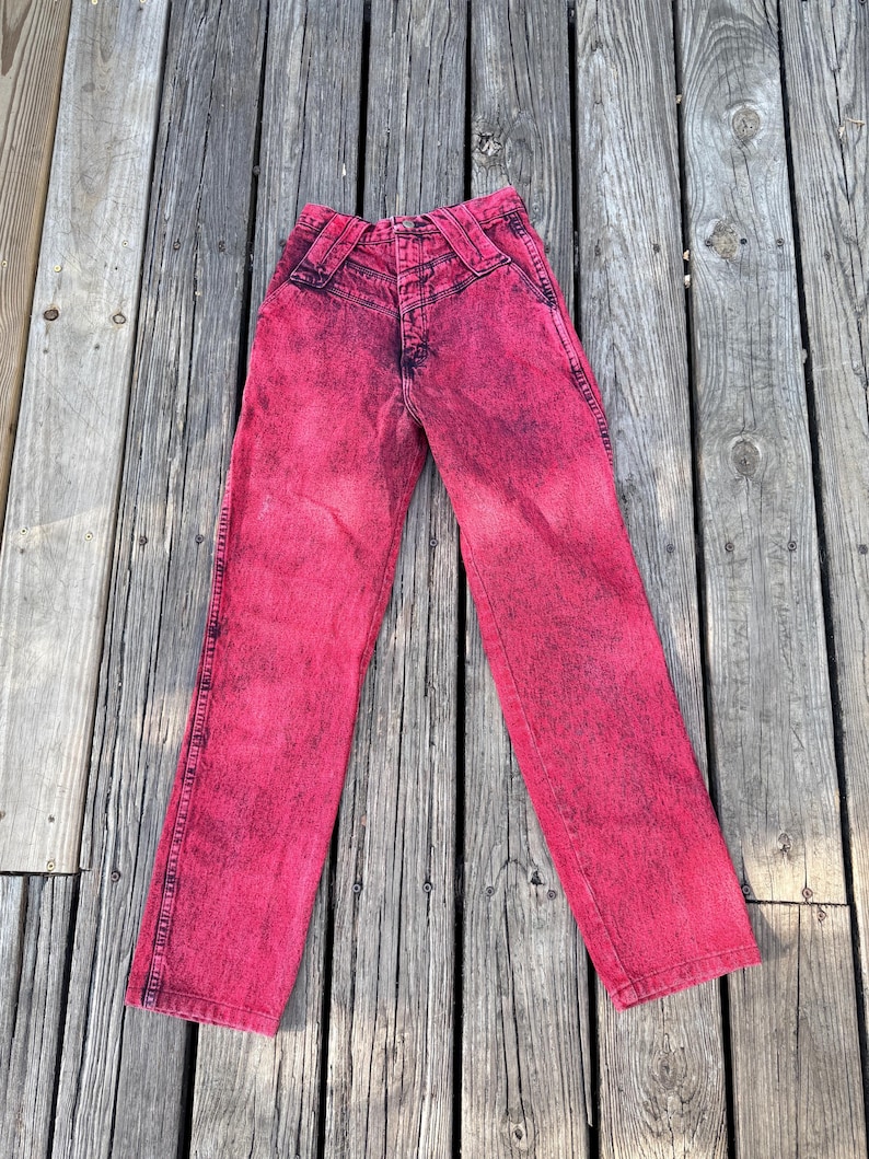 May include: A pair of acid wash pink denim jeans. The jeans have a high waist and a classic five-pocket design. The denim is a vibrant pink color with darker pink accents, giving it a unique, vintage look.
