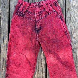 May include: Acid wash red jeans with a high waist and unique belt loops. The denim pants have a faded, distressed look with dark accents. A button and zipper closure is visible.