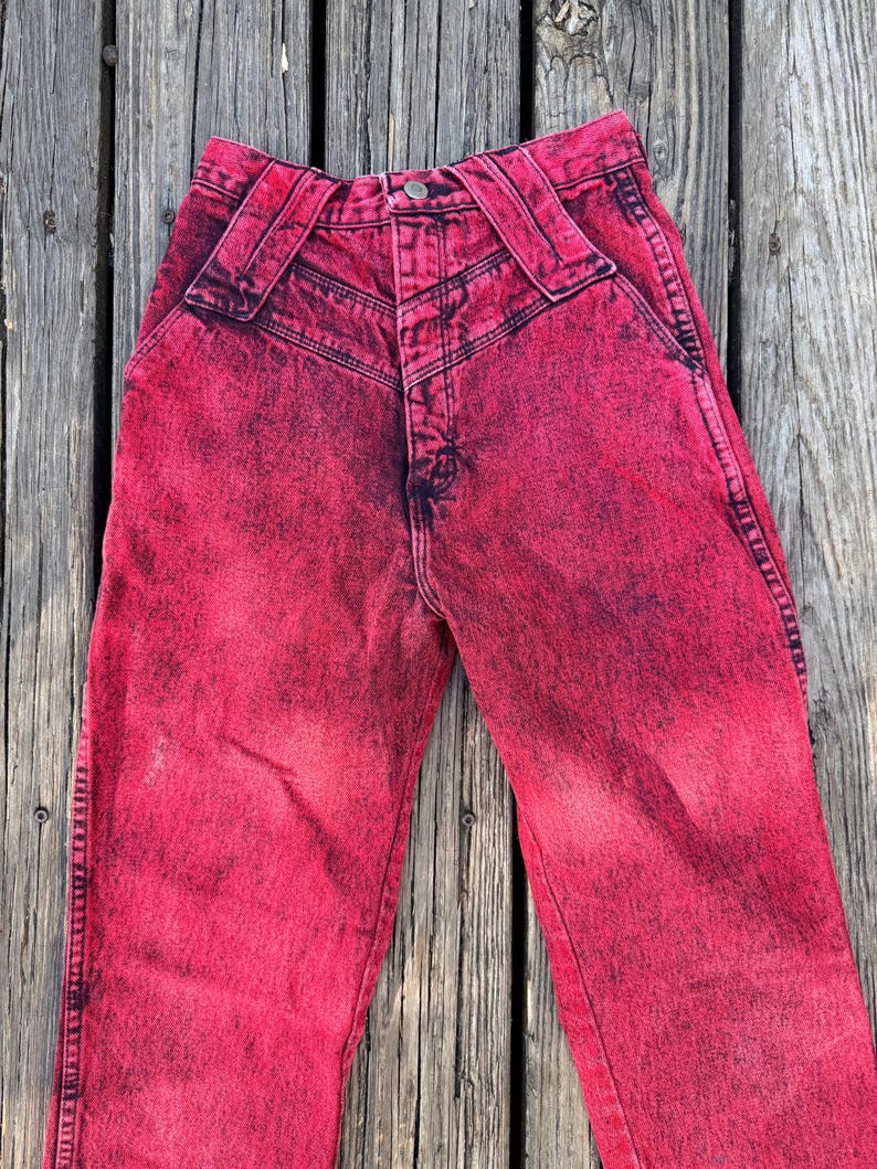 May include: Acid wash red jeans with a high waist and unique belt loops. The denim pants have a faded, distressed look with dark accents. A button and zipper closure is visible.