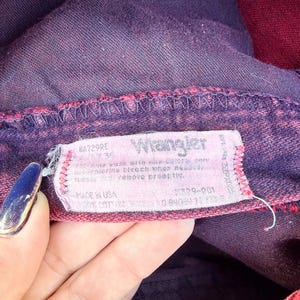 May include: Close-up of a pair of purple and red Wrangler jeans. The jeans' tag is visible, displaying the brand name and care instructions. The fabric appears to be a faded denim with a vintage aesthetic.