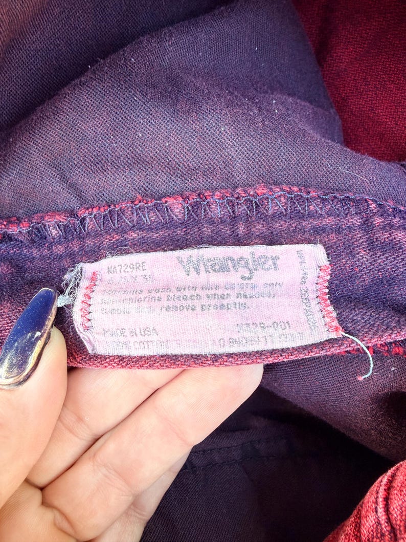 May include: Close-up of a pair of purple and red Wrangler jeans. The jeans' tag is visible, displaying the brand name and care instructions. The fabric appears to be a faded denim with a vintage aesthetic.