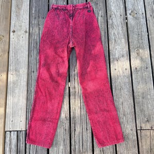 May include: A pair of acid-wash pink denim jeans. The jeans have a high waist and a straight leg cut. The denim has a faded, mottled appearance, with darker pink areas contrasting with lighter pink. The jeans are laid out on a weathered wooden surface.