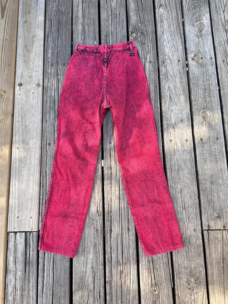 May include: A pair of acid-wash pink denim jeans. The jeans have a high waist and a straight leg cut. The denim has a faded, mottled appearance, with darker pink areas contrasting with lighter pink. The jeans are laid out on a weathered wooden surface.