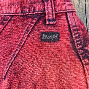 May include: Close-up of red acid wash denim jeans. The jeans feature a black Wrangler label and a belt loop. The denim has a textured appearance with dark red and black accents.