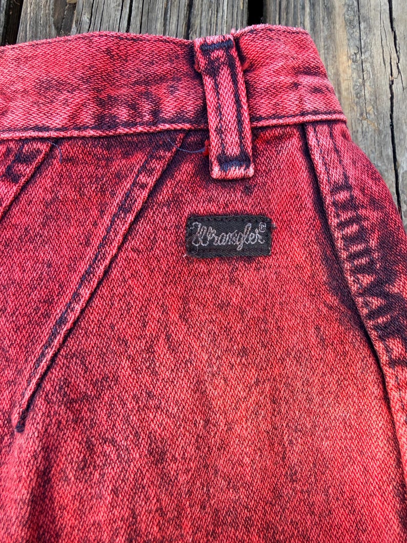 May include: Close-up of red acid wash denim jeans. The jeans feature a black Wrangler label and a belt loop. The denim has a textured appearance with dark red and black accents.
