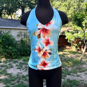 May include: A light blue sleeveless halter top featuring a vertical pattern of large white and red hibiscus flowers running down the center. The garment has a V-neckline and a fitted silhouette, displayed on a black mannequin torso.