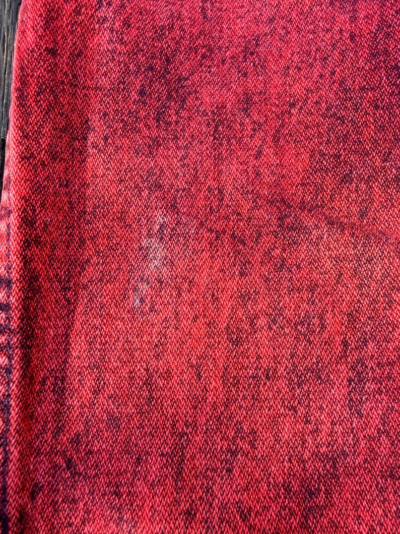 May include: Close-up of a textured fabric with a red and black speckled pattern. The fabric has a woven appearance, with the red color dominating and the black speckles creating a contrasting effect. The texture appears slightly rough.