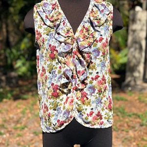 May include: A sleeveless floral print top with a V-neck and ruffled trim. The top features a white background with a pattern of red, purple, and green flowers. The top is displayed on a mannequin.
