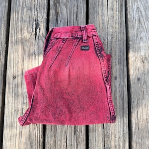 May include: Folded pair of red acid wash Wrangler jeans. The jeans have a black Wrangler label and stitching details. The jeans are folded neatly on a weathered wooden surface.