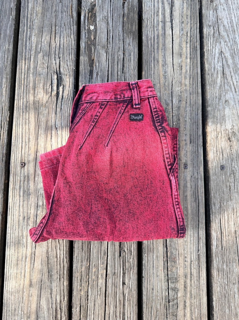 May include: Folded pair of red acid wash Wrangler jeans. The jeans have a black Wrangler label and stitching details. The jeans are folded neatly on a weathered wooden surface.