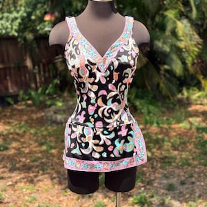 May include: A vintage black swimsuit with a colorful floral pattern. The one-piece suit features a V-neck and a skirted bottom with a pink border. The pattern includes pink, blue, orange, and white accents.