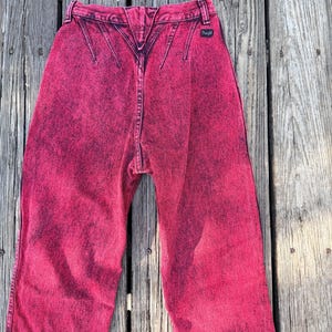 May include: Bright pink acid wash jeans with a high waist and a distinctive back yoke design. The jeans feature a classic five-pocket style and a small Wrangler label. The denim has a faded, distressed look.