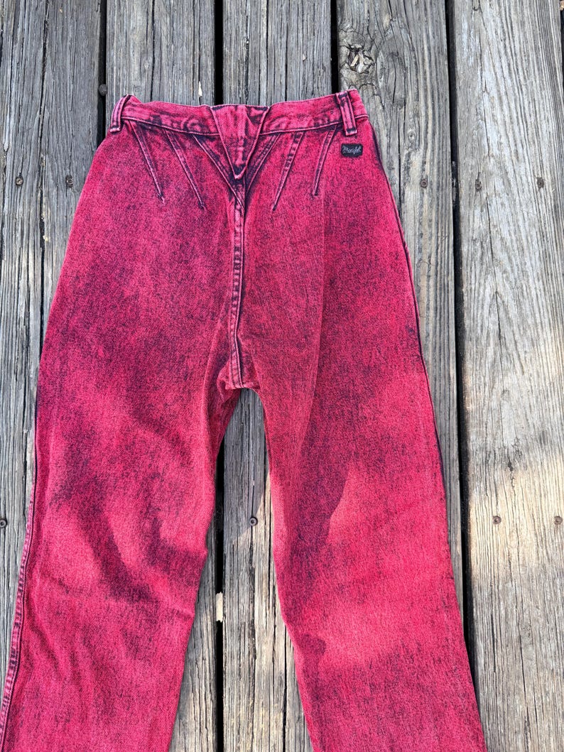May include: Bright pink acid wash jeans with a high waist and a distinctive back yoke design. The jeans feature a classic five-pocket style and a small Wrangler label. The denim has a faded, distressed look.
