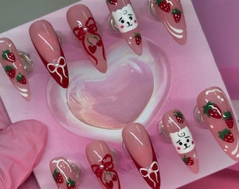 May include: A set of ten almond-shaped press-on nails featuring a nude base with hand-painted red strawberries, white bows, and a white cartoon cat face. The designs include red French tips with white bows and scattered red strawberry accents.