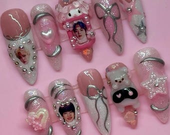 May include: A set of ten long, pointed artificial nails. The nails are primarily pink and white, with silver accents, glitter, and various decorative elements, including bows, hearts, and character figurines. Some nails feature images of people.
