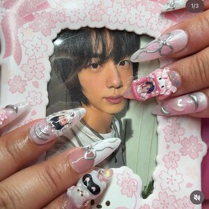 Puede incluir: U&ntilde;as largas en forma de almendra, en tonos rosa, blanco y plateado. Las u&ntilde;as presentan purpurina, lazos y peque&ntilde;os dise&ntilde;os de personajes. Una foto enmarcada de una persona est&aacute; en el fondo.