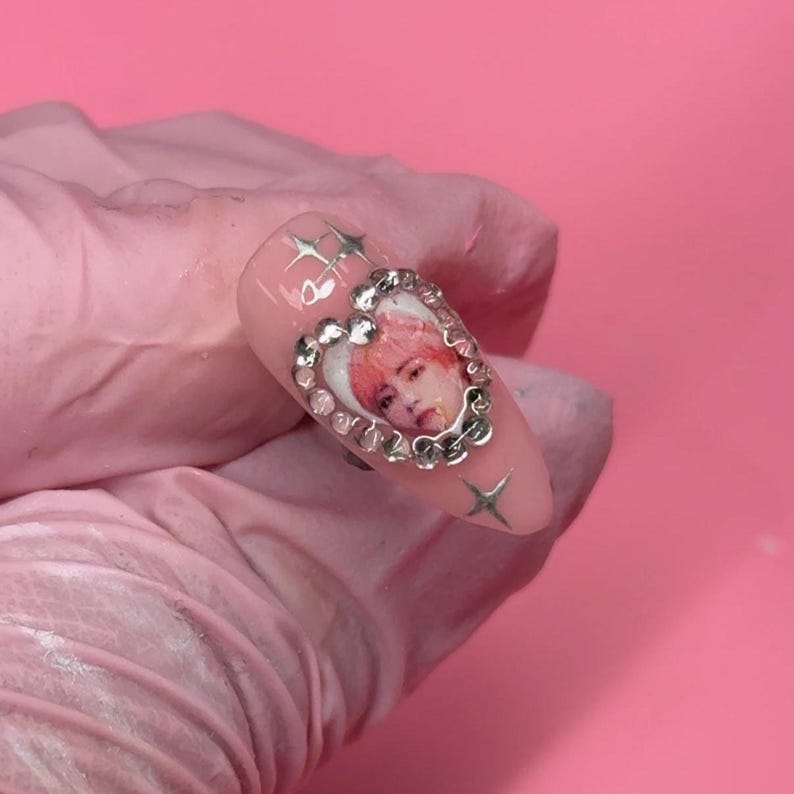 Puede incluir: Una u&ntilde;a rosa con un dise&ntilde;o en forma de coraz&oacute;n que presenta un retrato de una persona con cabello rosado, rodeado de pedrer&iacute;a. Tambi&eacute;n hay adornos plateados en forma de estrella.