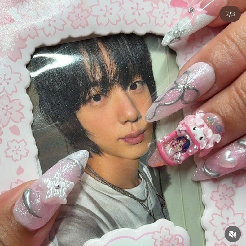 Puede incluir: Primer plano de una mano con u&ntilde;as acr&iacute;licas largas y rosas. Las u&ntilde;as presentan detalles plateados, lazos y un dise&ntilde;o 3D de Hello Kitty. En el fondo, una foto enmarcada de una persona.