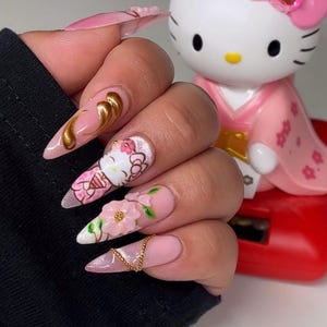 Puede incluir: Uñas acrílicas largas y puntiagudas con base rosa y varios diseños. Una uña presenta un remolino dorado, otra un diseño de Hello Kitty y una tercera tiene una flor en 3D. Una cadena dorada y detalles de purpurina completan el look. Una figura de Hello Kitty está en el fondo.