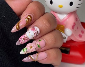 May include: Long, pointed acrylic nails with a pink base and various designs. One nail features a gold swirl, another a Hello Kitty design, and a third has a 3D flower. A gold chain and glitter accents complete the look. A Hello Kitty figurine is in the background.