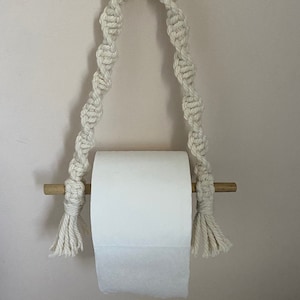 May include: A white macrame toilet paper holder with a wooden dowel and a white wooden ring. The holder is hanging from a hook on a wall.