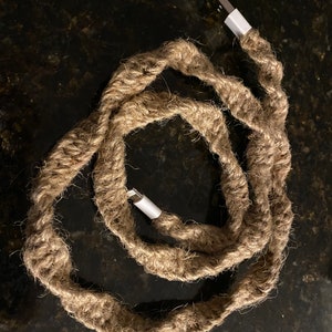 May include: A USB charging cable wrapped in natural-colored rope. The cable has white connectors at each end. The rope is tightly wound around the cable, creating a textured, rustic appearance. The cable is coiled.