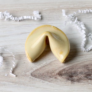 May include: A ceramic fortune cookie in a light yellow color. The cookie is in the shape of a half-moon with a crease in the middle.
