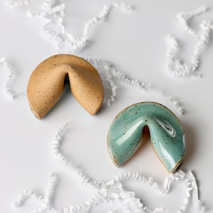 May include: Two ceramic fortune cookie ornaments, one is speckled brown and the other is speckled light blue.