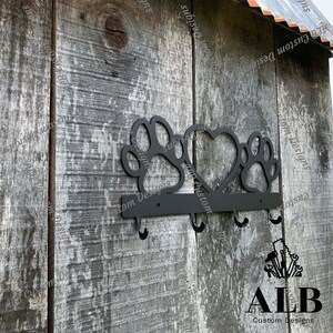 Pu&ograve; includere: Portachiavi in metallo nero con tre ganci a forma di impronte di zampe e un cuore. Il portachiavi &egrave; montato su una parete di legno con il testo "ALB Custom Designs" sotto.