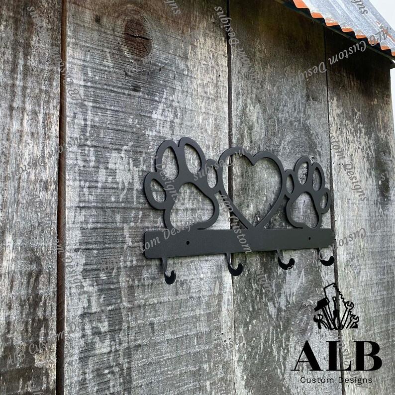 Pu&ograve; includere: Portachiavi in metallo nero con tre ganci a forma di impronte di zampe e un cuore. Il portachiavi &egrave; montato su una parete di legno con il testo "ALB Custom Designs" sotto.
