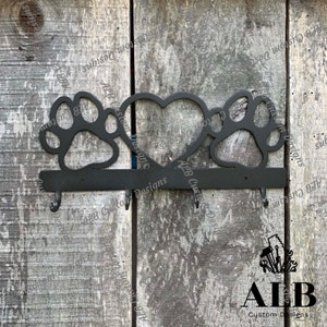 Pu&ograve; includere: Portachiavi in metallo nero con due impronte di zampa e un cuore al centro. Il portachiavi ha tre ganci e il testo "ALB Custom Designs" &egrave; inciso sul fondo.