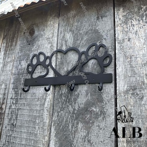 Pu&ograve; includere: Portachiavi in metallo nero con tre ganci. Il portachiavi ha la forma di un cuore con impronte di zampe su entrambi i lati. Il testo "ALB Custom Designs" &egrave; visibile sul portachiavi.