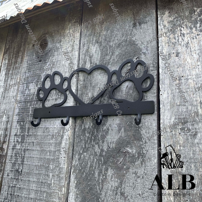 Pu&ograve; includere: Portachiavi in metallo nero con tre ganci. Il portachiavi ha la forma di un cuore con impronte di zampe su entrambi i lati. Il testo "ALB Custom Designs" &egrave; visibile sul portachiavi.