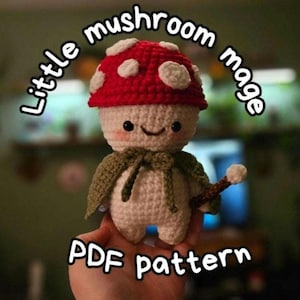 Puede incluir: Muñeca de mago de seta de ganchillo, con gorro rojo con lunares blancos, capa verde y varita de madera. La muñeca tiene una cara sonriente. El texto en la imagen dice "Little mushroom mage PDF pattern."
