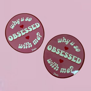 以下が含まれることがあります： 「Why u so obsessed with me?」という白い文字が書かれたピンク色の丸いステッカー2枚。 「so」と「obsessed」の間、「with」と「me」の間にハートマークがあります。 文字はレトロなフォントスタイルです。 ステッカーの下には白い文字で「Raiz」と書いてあります。