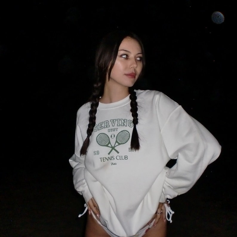 May include: A white sweatshirt with a green tennis club logo featuring two crossed tennis rackets and the text "SERVING CLUB" and "TENNIS CLUB Paris". The model is wearing the sweatshirt with their hair in two braids.