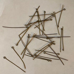 May include: A pile of gold-colored head pins with loops on one end. The pins are scattered on a white surface.