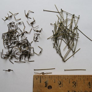 May include: A pile of gold-colored metal jump rings and a pile of gold-colored metal straight pins. A wooden ruler with inch markings is below the piles.