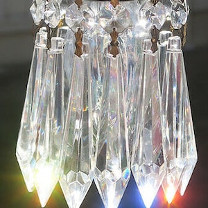 May include: Close-up of a crystal chandelier with multiple clear, faceted crystal pendants. The chandelier has a brass-colored metal frame and is illuminated with colorful lights, creating a sparkling effect.