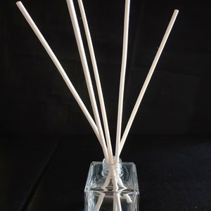 May include: A clear glass bottle with a square base and a narrow neck. Six white wooden sticks are inserted into the bottle. The bottle and sticks are on a black surface.