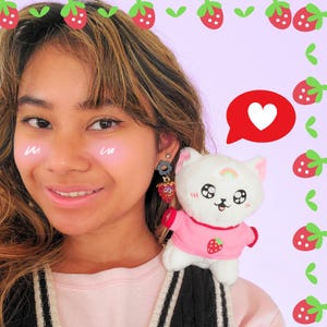 May include: A person with strawberry-themed accessories, including earrings and a plush toy. The earrings feature a flower and a strawberry charm. The plush toy is a white cat wearing a pink shirt with a strawberry graphic. A red speech bubble with a white heart is also visible.