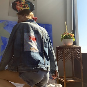 May include: A denim jacket with a patch featuring a red and white design and the word "Marlboro" in gold lettering. The jacket is worn with denim shorts. A sign that says "Caroline's Cafe" is visible in the background.