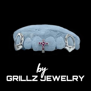 May include: Silver grillz with a cross design featuring pink gemstones. The grillz are displayed on a blue mold. The text "by GRILLZ JEWELRY" is displayed below the grillz.