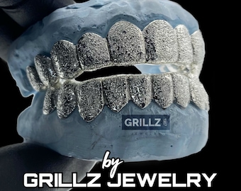 Może przedstawiać: Zbliżenie srebrnej grillz z fakturą przypominającą diament. Grillz są umieszczone w formie ust. Tekst "GRILLZ JEWELRY" jest widoczny na dole obrazu.
