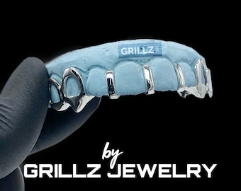 May include: Silver grillz with a blue mold, featuring a unique design with multiple teeth. The grillz are labeled "GRILLZ by GRILLZ JEWELRY".