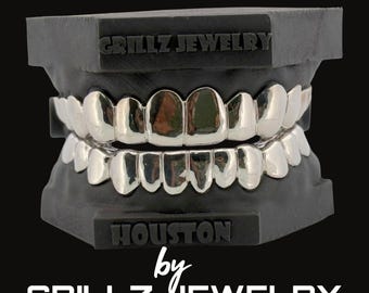 Może przedstawiać: Srebrne grillzy prezentowane na czarnym stojaku. Na górze stojaka widnieje napis "GRILLZ JEWELRY", a na dole "HOUSTON by GRILLZ JEWELRY."
