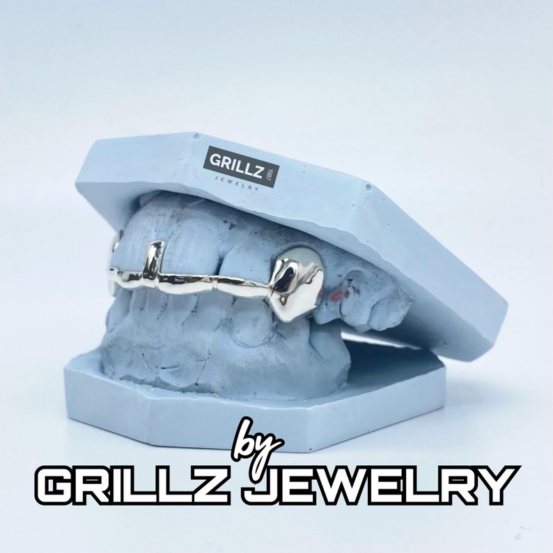 K&ouml;nnte beinhalten: Ein silberner Grillz f&uuml;r Z&auml;hne, auf einem blauen Zahnmodell ausgestellt. Der Grillz ist einteilig mit einem geschwungenen Design. Das Modell ist mit "Grillz Jewelry" beschriftet.
