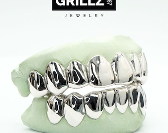 May include: Silver grillz with a shiny finish, set in a mould of a mouth. The grillz are made of multiple pieces that fit together to cover the front teeth. The brand name "GRILLZ 1987 JEWELRY" is visible at the top of the image.